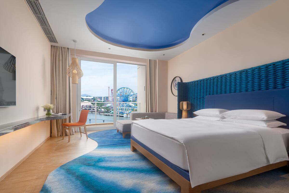 A modern hotel room with a large bed, a desk with a chair, a view of a Ferris wheel, and a balcony.