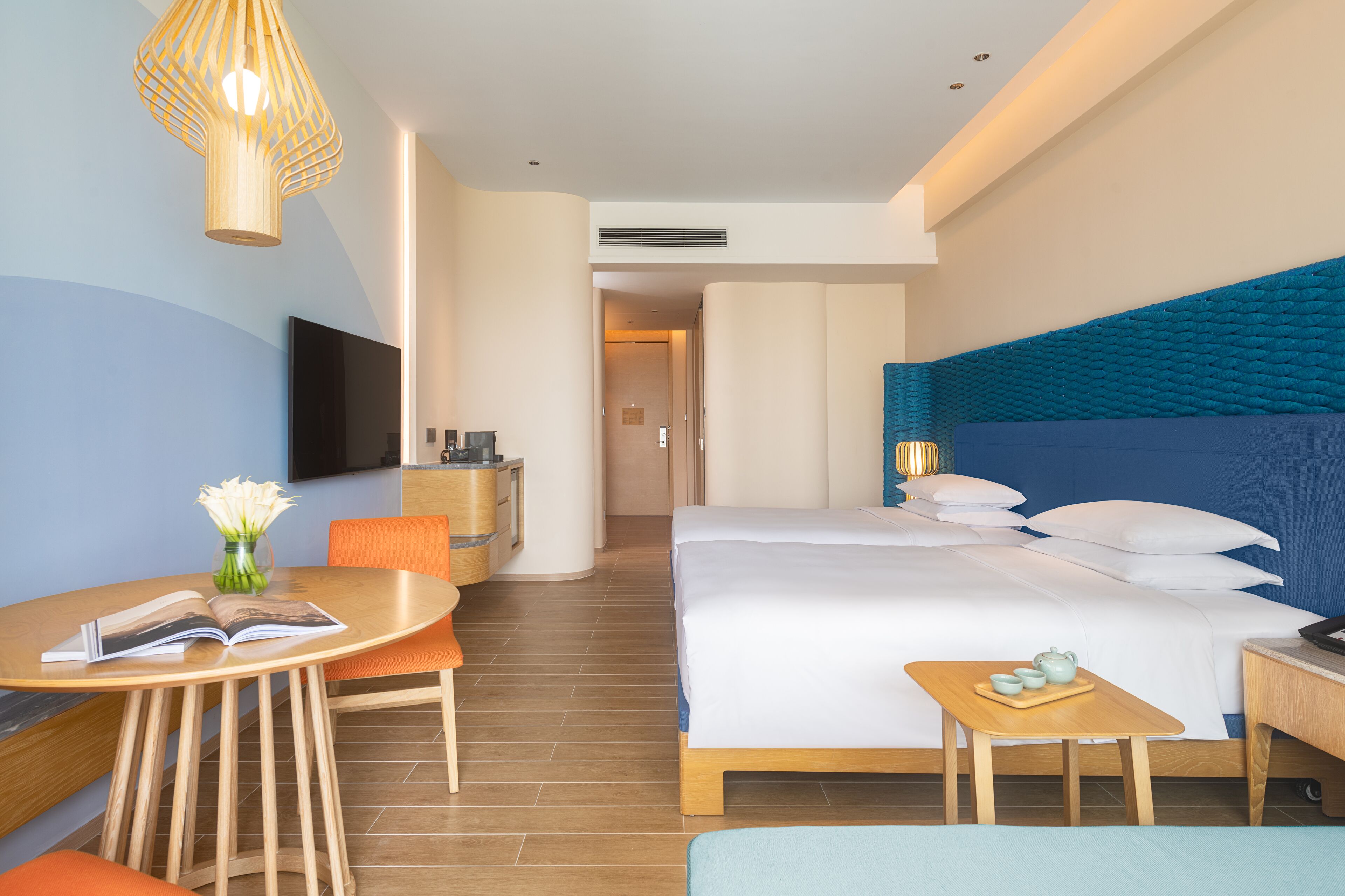 A hotel room with a bed, a desk, a chair, a TV, and a bedside table with a lamp.