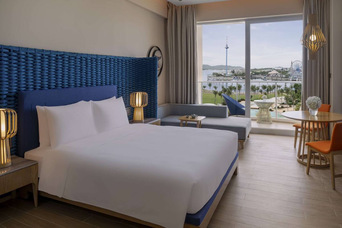 A hotel room with a large bed, a sofa, a dining table with chairs, and a view of a body of water and a tower.