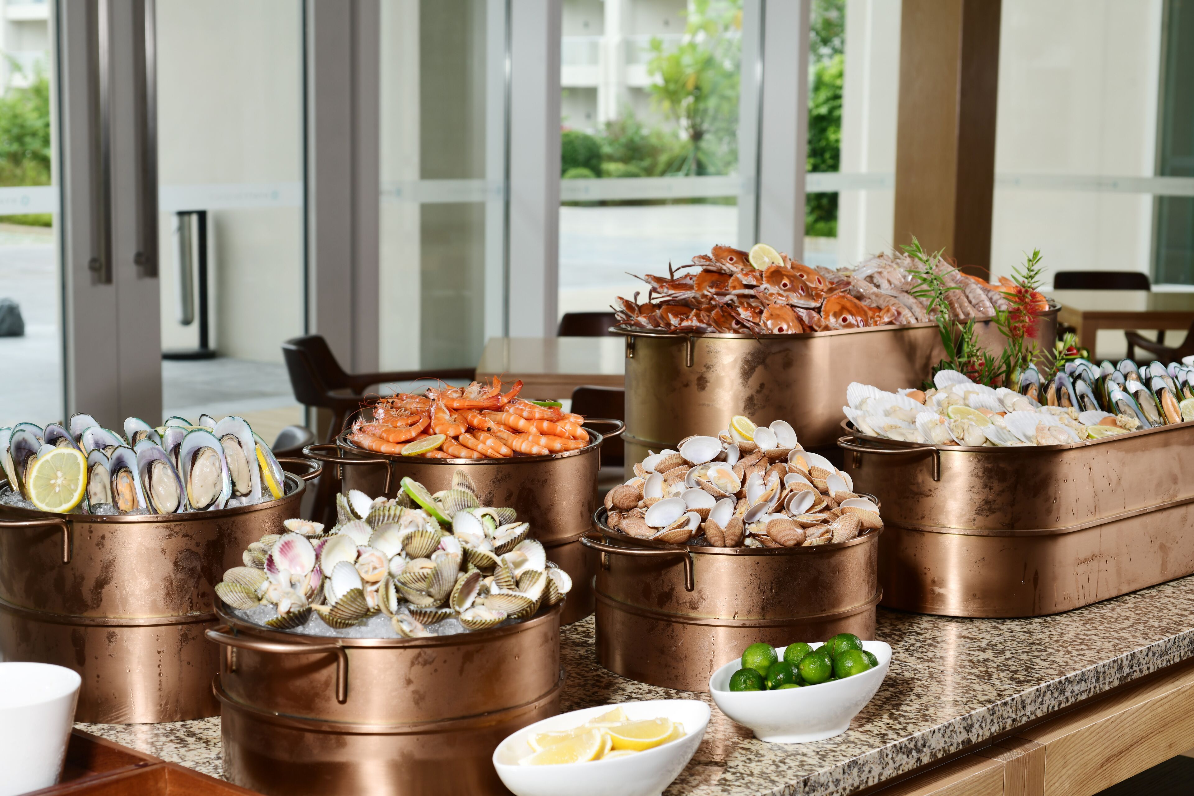 A buffet with various seafood options in copper pots, a bowl of lime wedges, and a view of the outdoor area.