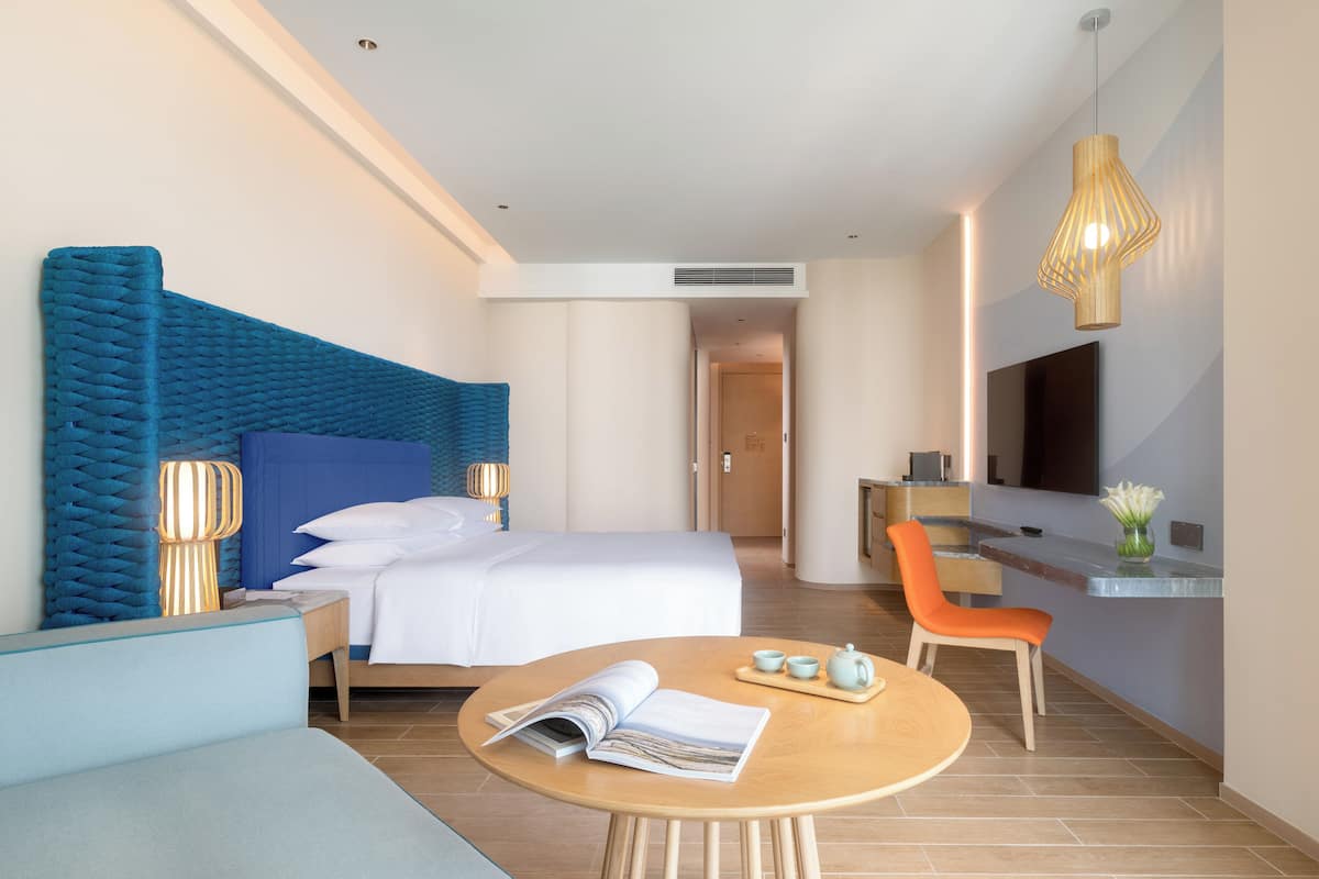 A modern hotel room with a large bed, a desk, a TV, and a seating area.