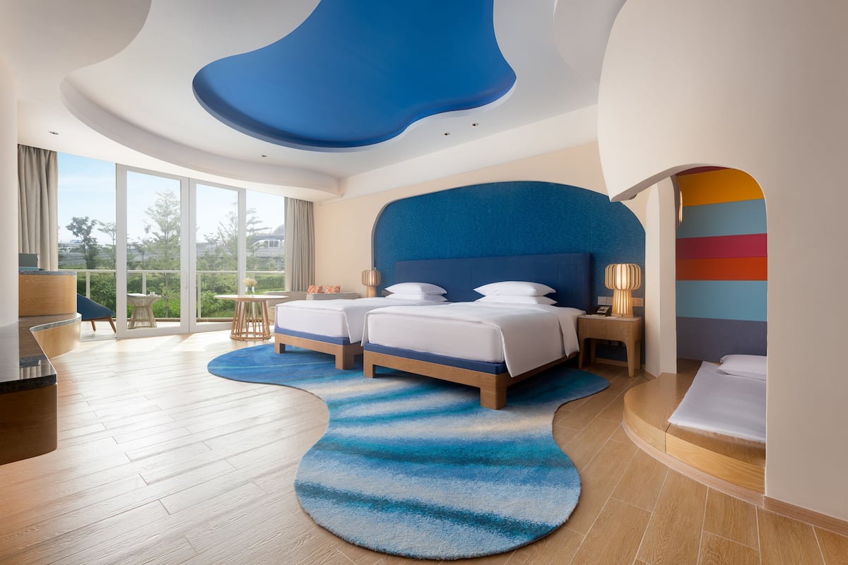 A modern hotel room with two beds, a large window, and a view of the outdoors.