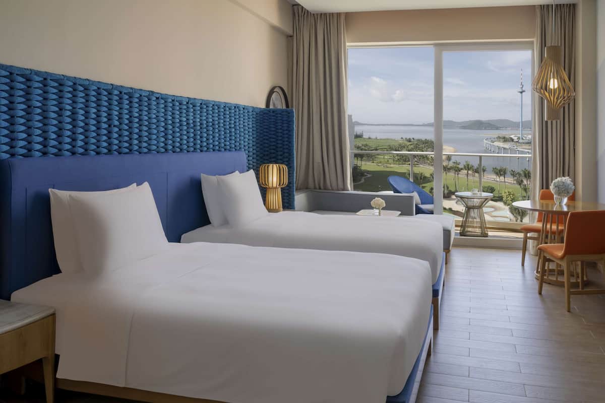 A hotel room with two beds, a large window with a view of a body of water and a bridge, and a balcony with a table and chairs.