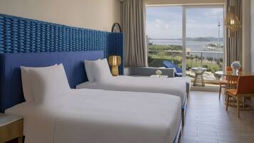 Habitación de hotel con dos camas, un ventanal con vista a un cuerpo de agua y un puente, y un balcón con mesa y sillas.
