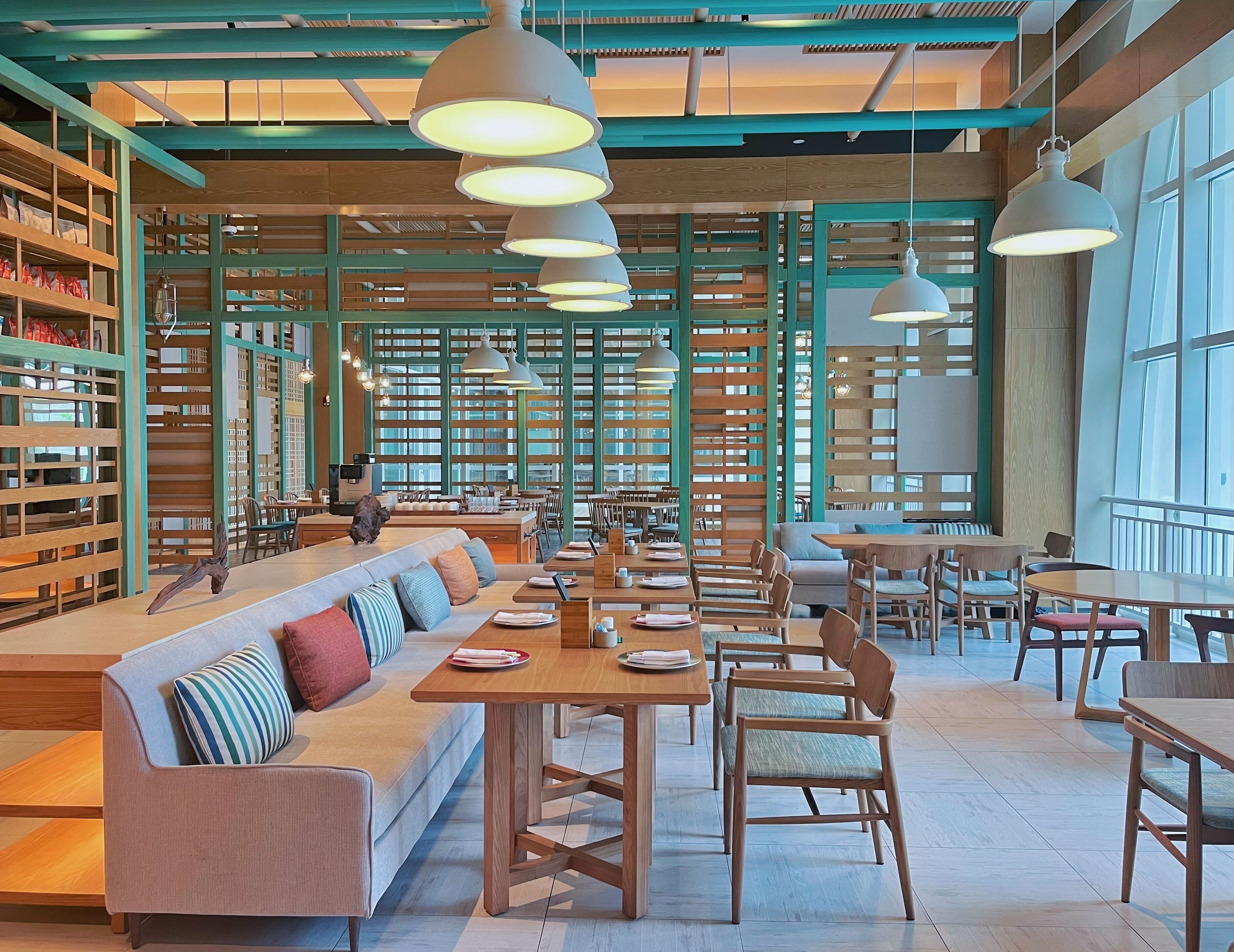 A spacious cafe with wooden furniture, large windows, and hanging pendant lights.