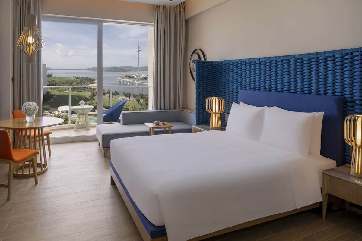 A hotel room with a large bed, a sofa, a small table, and a chair. There is a view of a body of water and a tower in the distance.