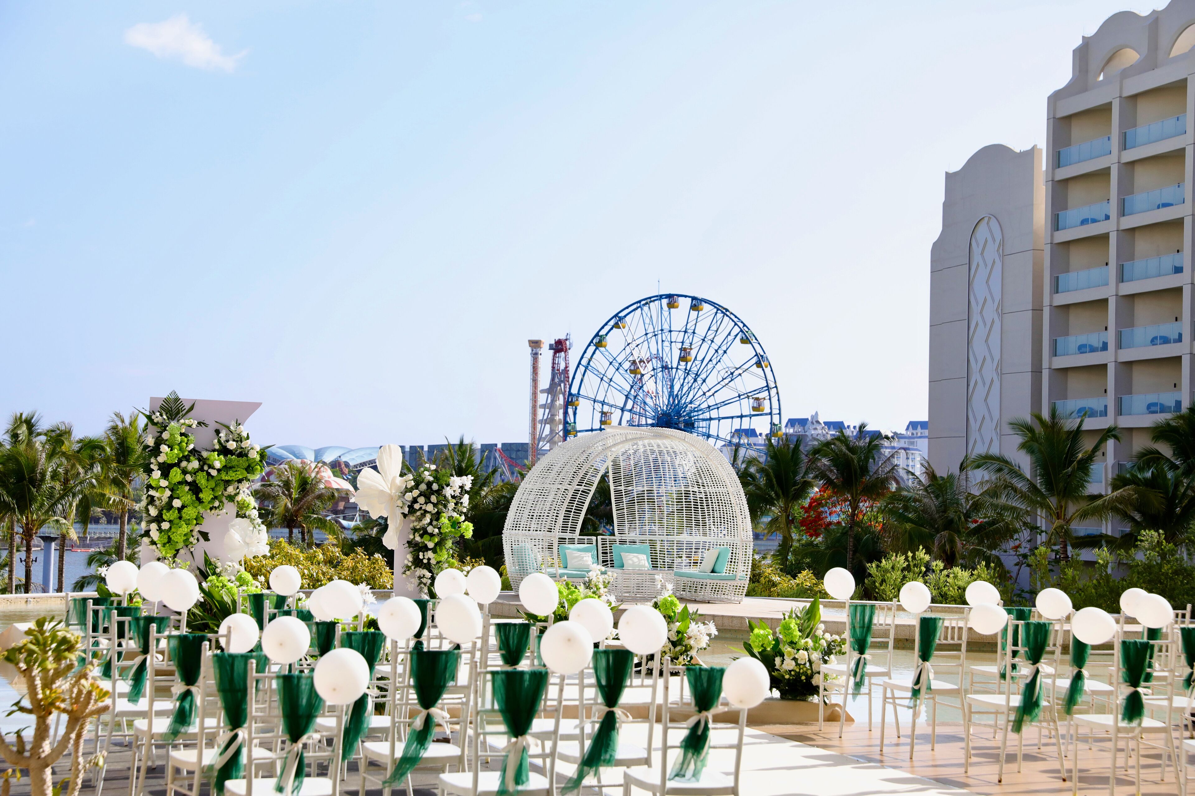 A beachside event setup with white chairs, a large white dome structure, and a Ferris wheel in the background.