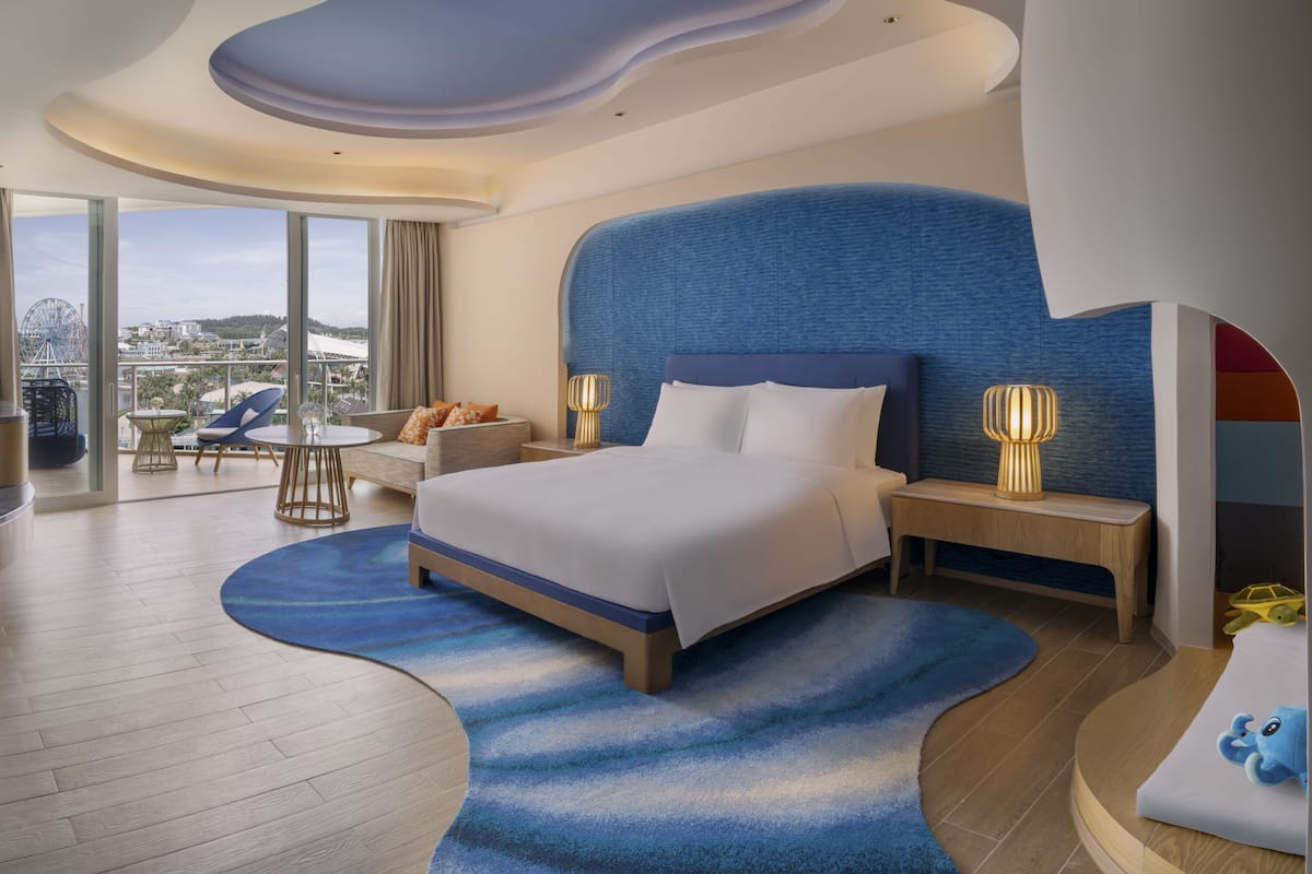 A modern hotel room with a large bed, a curved blue rug, a wooden floor, a view of a cityscape, and a balcony with a chair and a table.