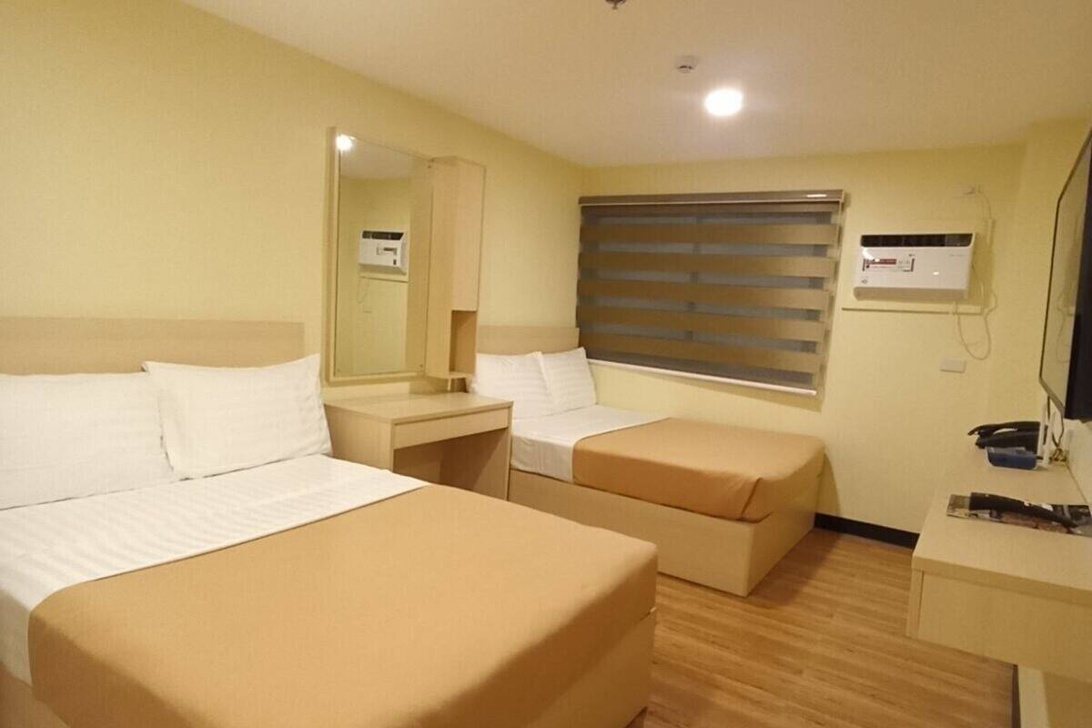 A hotel room with two beds, a desk, a chair, a window with blinds, and an air conditioning unit.