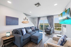 Een woonkamer met een grijze bank, een kleine tafel en een stoel. Er is een raam met uitzicht op zee en een gebouw.