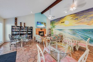 A living room with a fireplace, a large bookshelf, a dining area with white chairs, and a mural of a sunset over the ocean.