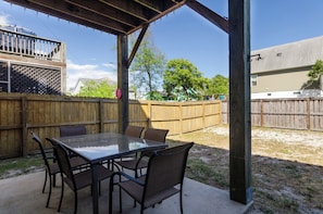 A covered patio with a glass table and chairs, a wooden fence, and a house in the background.