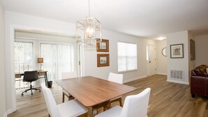A spacious dining area with a wooden table and white chairs, a modern pendant light, and a view of a kitchen and dining area through large windows. - 19 F Pinehurst Manor: 1  BR, 1  BA Condominium in Pinehurst, Sleeps 3 (Pinehurst)