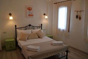 Uma cama bem arrumada com lençóis brancos e manta bege, um criado-mudo verde com gavetas e uma luminária fixada na parede.