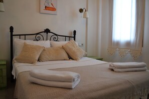 Uma cama bem arrumada com colcha estampada e travesseiros, uma luminária na parede e uma janela com cortinas.