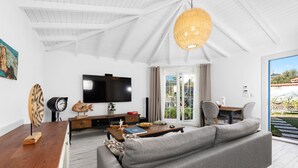 A modern living room with a large gray sofa, wooden coffee table, and a flat-screen TV. There is a wicker chandelier, a painting on the wall, and a view of the outdoors through the sliding glass door. - Rhodium Hotel (Cesme)