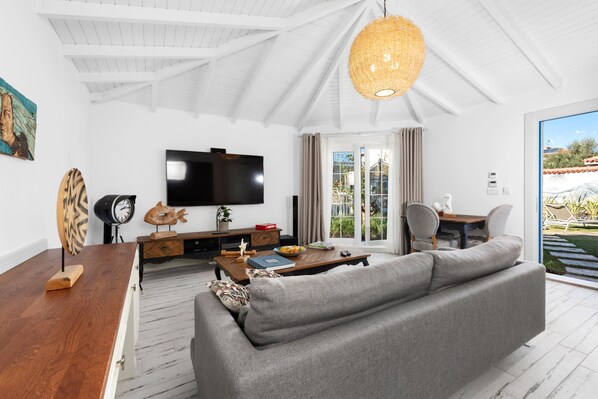 A modern living room with a large gray sofa, wooden coffee table, and a flat-screen TV. There is a wicker chandelier, a painting on the wall, and a view of the outdoors through the sliding glass door. - Rhodium Hotel (Cesme)