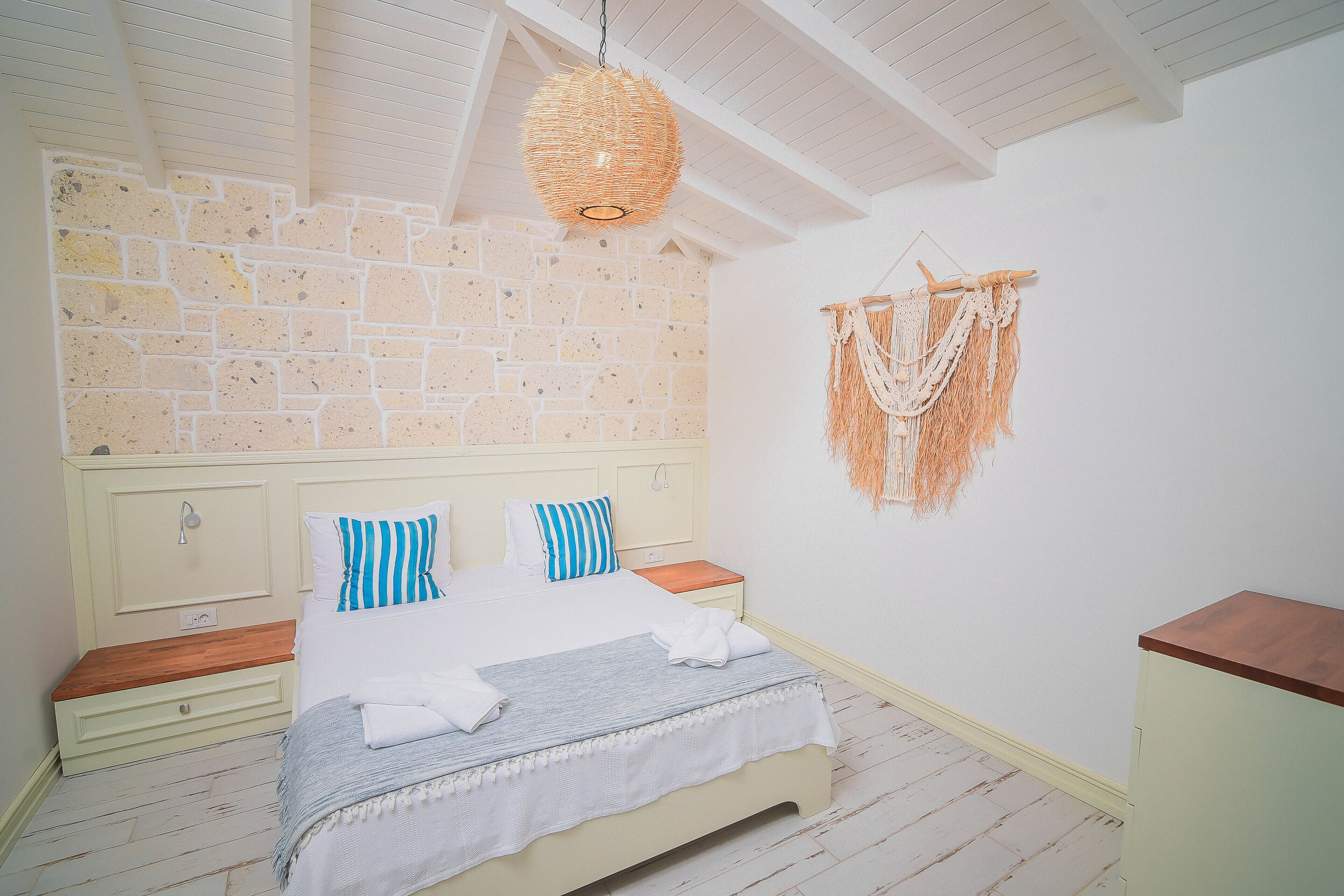 A bedroom with a brick wall, a bed with blue and white striped pillows, a woven hanging light fixture, and a macrame wall hanging.