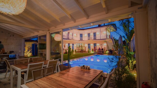 An indoor pool area with wooden tables and chairs, a pool, and a view of a multi-story building with balconies. - Rhodium Hotel (Cesme)