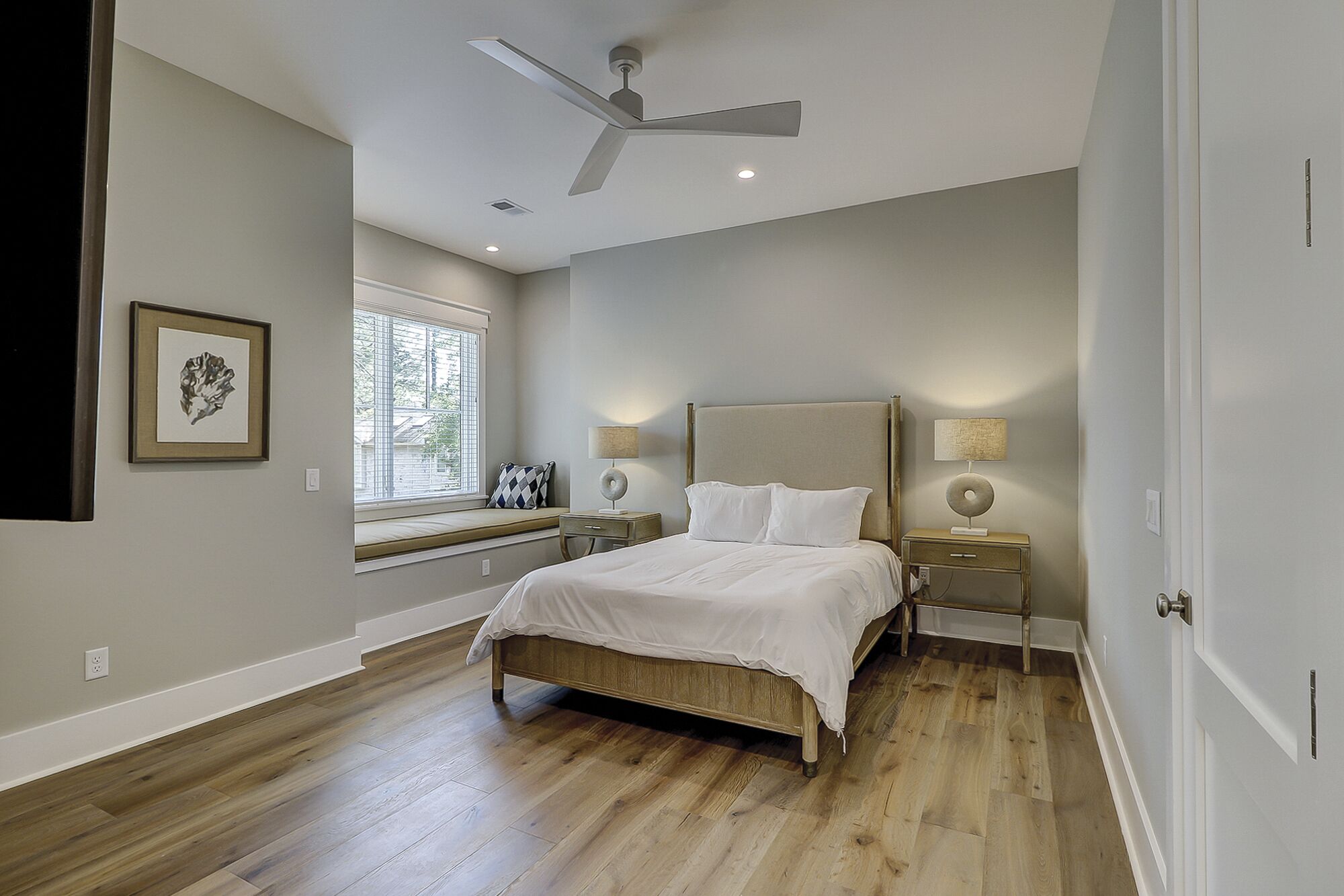 Une chambre à coucher comprenant un lit, des tables de chevet, une fenêtre avec des stores, un ventilateur de plafond et un luminaire fixé au mur.