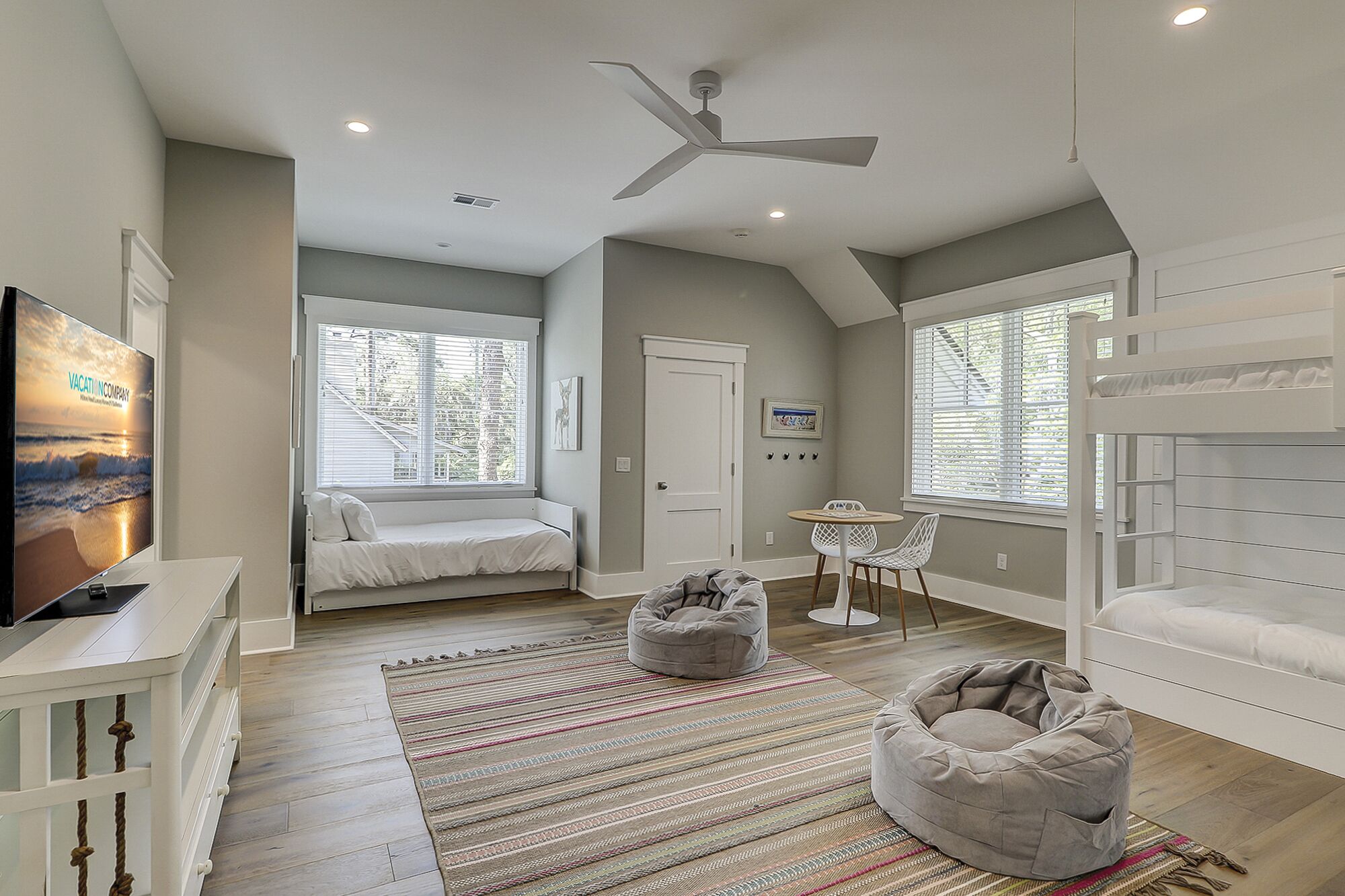 A spacious room with a large bed, a desk, a chair, and a bunk bed. There is a ceiling fan and a window with blinds.
