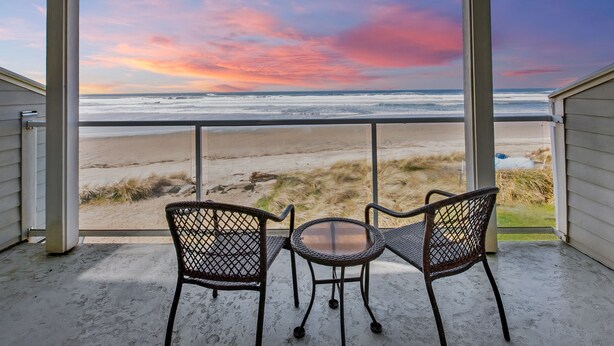A balcony with two wicker chairs and a small table, overlooking a beach with a sunset view.