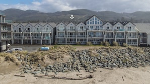 A coastal hotel with multiple balconies, a rocky beach in the foreground, and mountains in the background.
