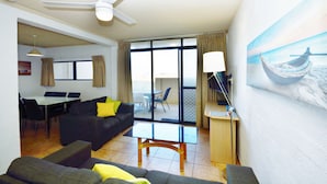 Quarto de hotel com varanda, área de refeições, sala de estar com sofá e mesa de centro, e um quadro de um barco na parede.
