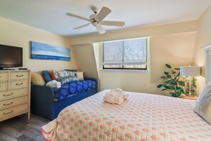 Un dormitorio con cama, ventilador de techo, televisor, cómoda, ventana con persianas y un cuadro en la pared.