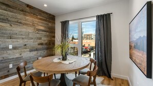 A dining area with a round wooden table, four chairs, a vase with plants, and a large window with curtains. - Rustic Overlook- 30 day rental in the new Merin building in downtown Bozeman! (Bozeman)