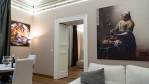 Una sala de estar con un sofá blanco, un comedor con una mesa preparada para dos personas, y un cuadro grande en la pared.
