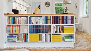 Ein ordentlich bestelltes Bücherregal mit vielen Büchern, einer Uhr und einem Fenster mit Grünpflanzen draußen.