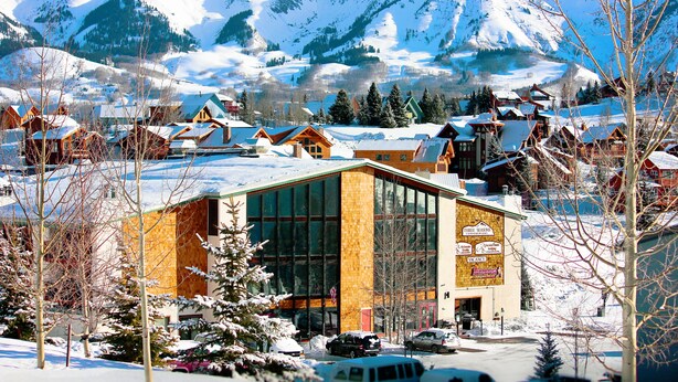 A modern building with large windows, surrounded by snow-covered houses and mountains in the background.