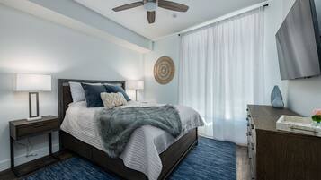 Un dormitorio con cama, mesitas de noche, escritorio, ventilador de techo y un amplio ventanal con cortinas.