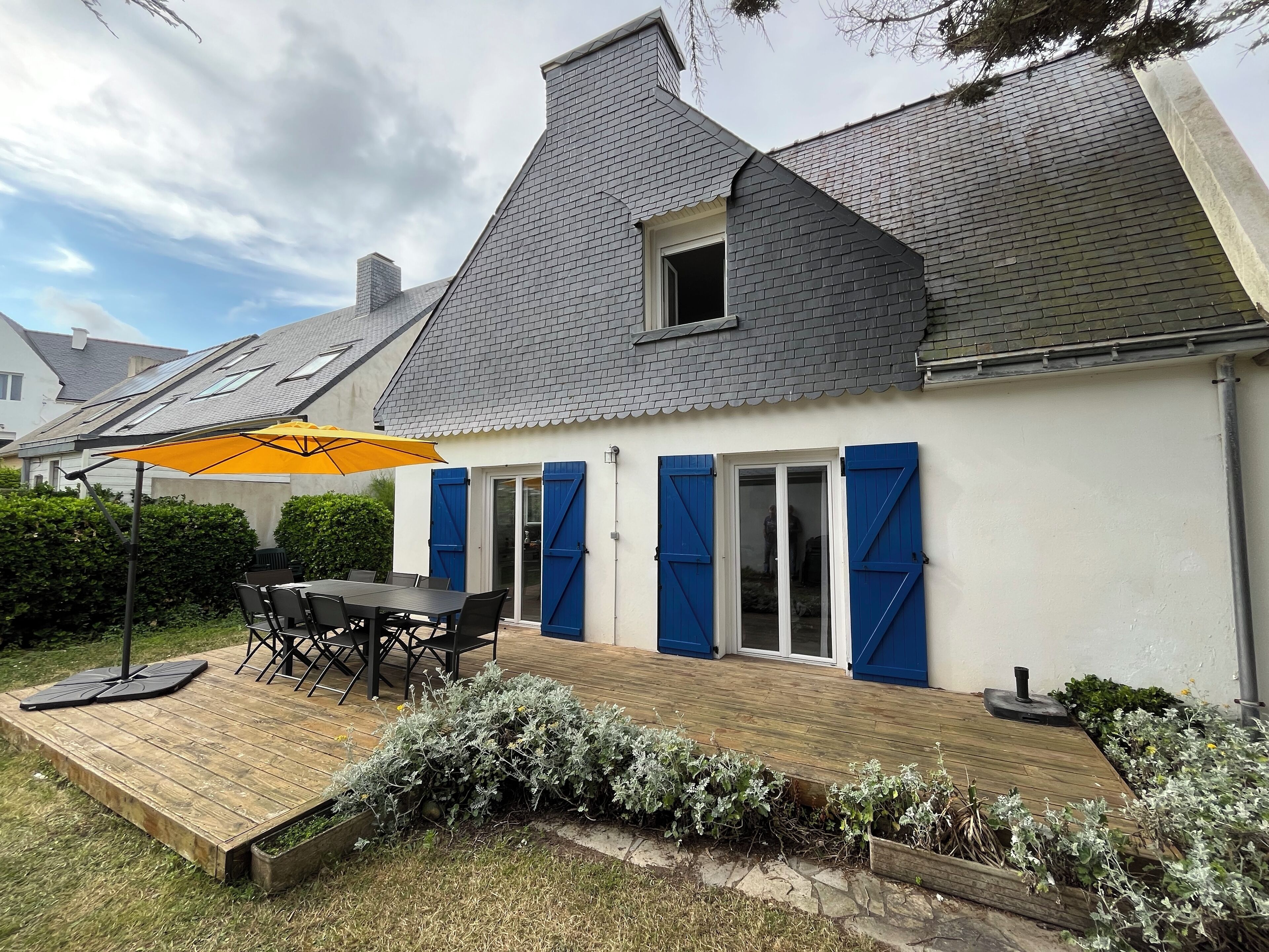 Une maison avec une terrasse en bois, une table avec des chaises et un parasol jaune.