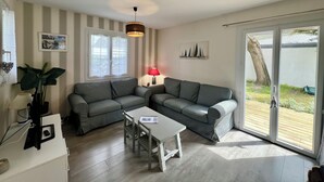 Una sala de estar con un sofá seccional gris, una mesa de centro blanca y una lámpara roja. Las paredes tienen rayas, hay una lámpara en el techo y una ventana con persianas.