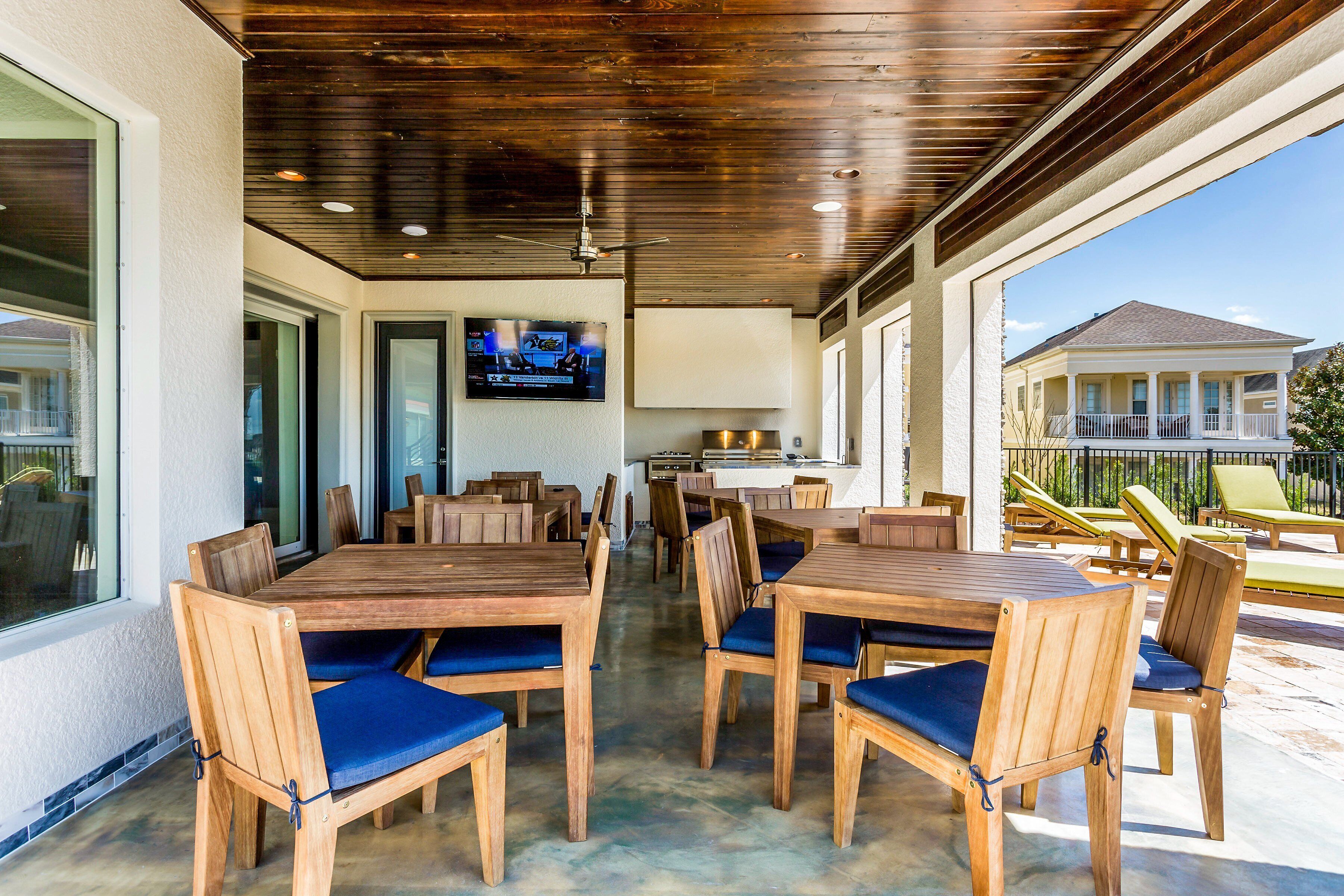 An outdoor dining area with wooden tables and chairs, a TV mounted on the wall, and a view of a house in the background.