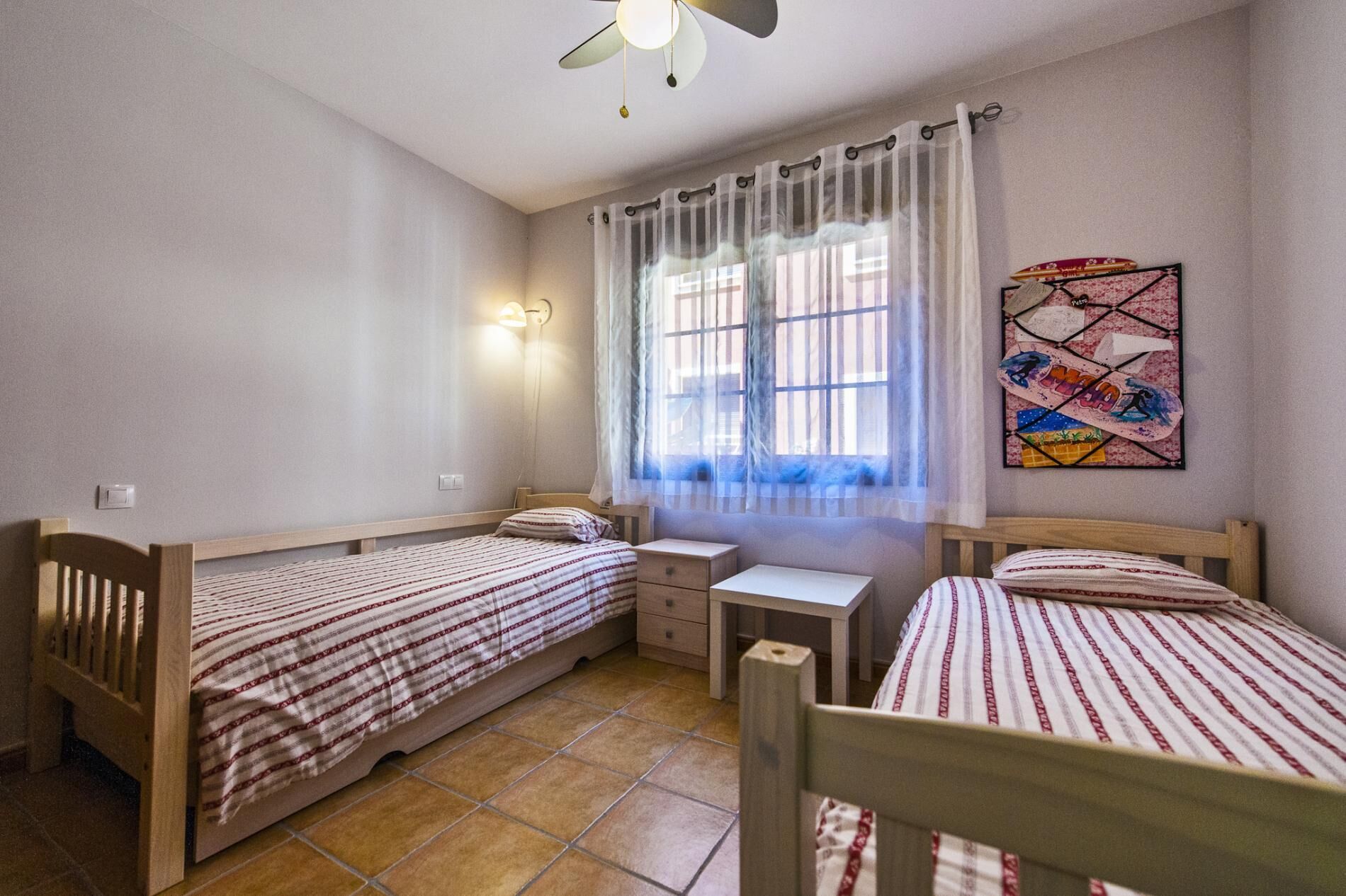 Ein Schlafzimmer mit zwei Holzbetten, einem kleinen Nachttisch, einem Hocker, einem Deckenventilator und einem Fenster mit durchsichtigen Vorhängen.