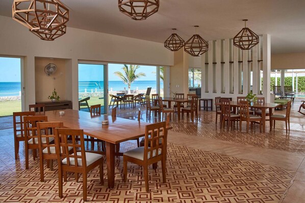 A spacious dining area with a large wooden table and chairs, large windows offering a view of the beach, and geometric patterned flooring. - Stunning beachfront Suites Wi-Fi East of Progreso (Chicxulub Puerto)