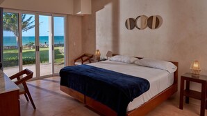 Ein Schlafzimmer mit einem großen Bett, einem Schreibtisch und einem Stuhl. Durch die Schiebetüren ist der Blick aufs Meer.