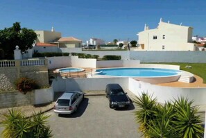 A residential property with a swimming pool, two cars parked in the driveway, and a white building with a red roof.
