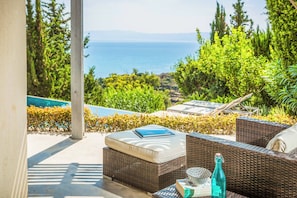 A wicker outdoor seating area with a view of the sea, a pool, and lush greenery.