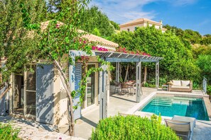 A poolside area with a pergola, outdoor seating, and a view of a hillside house.