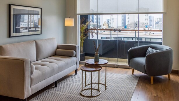 A modern living room with a sofa, armchair, and a small table with a vase. A large window offers a view of a cityscape.