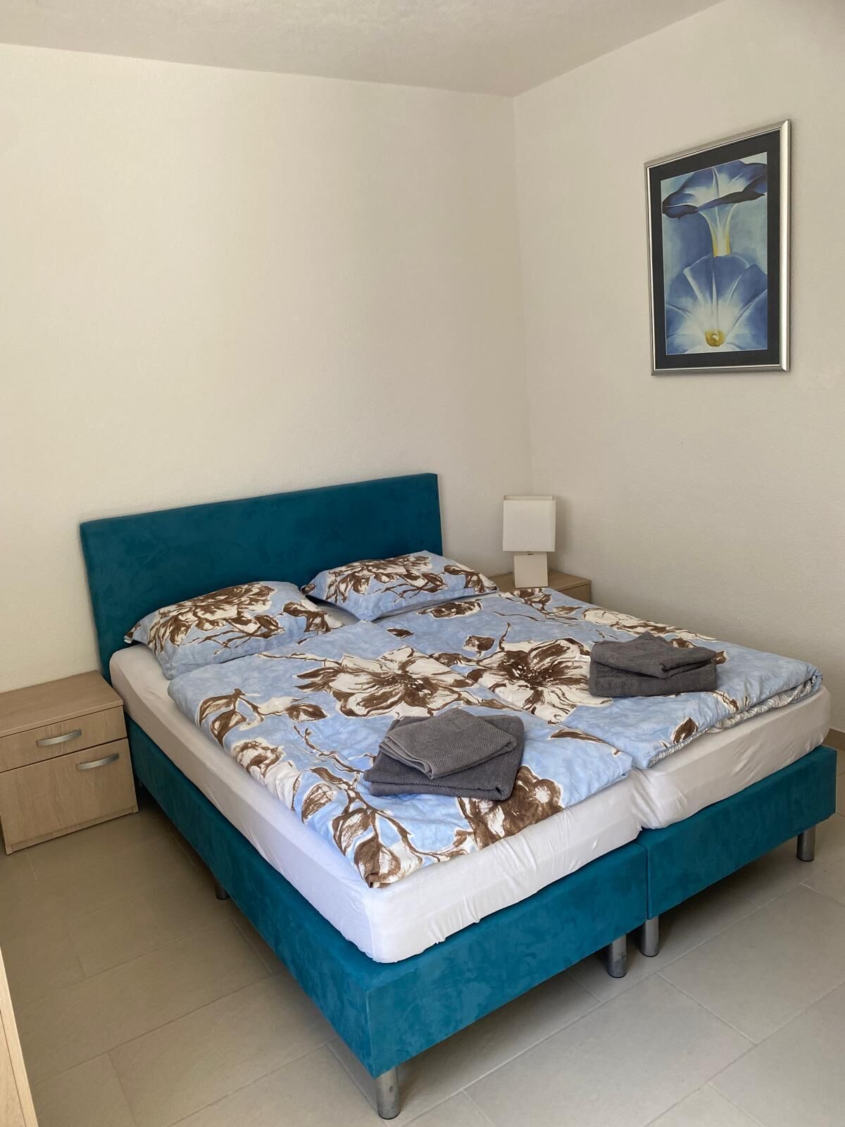 Een net opgemaakt bed met een bloemenbedekker, een blauw hoofdbord en een nachtkastje met een lamp.
