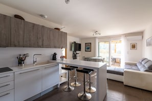 Una cocina moderna con armarios blancos, una isla central con taburetes de bar, un comedor con mesa y sillas, y una sala de estar con sofá.