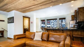 Een woonkamer met een bruine leren hoekbank, een houten plafond en een keuken met houten kastjes en een roestvrijstalen koelkast.