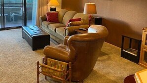 A living room with a sofa, armchair, and ottoman, a balcony with a view, and two wall-mounted lamps. - Updated Condo Just a Short Walk to the Slopes (Whitefish)