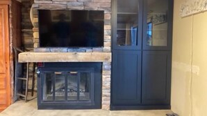 A living room with a stone fireplace, a flat-screen TV mounted above it, and a dark-colored cabinet to the right. - Updated Condo Just a Short Walk to the Slopes (Whitefish)