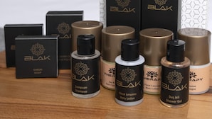 A selection of BLAK personal care products on a wooden surface, including shampoo, body lotion, and shower gel, with a bed and nightstand in the background. - Depiero Hotel Karaköy (Istanbul)